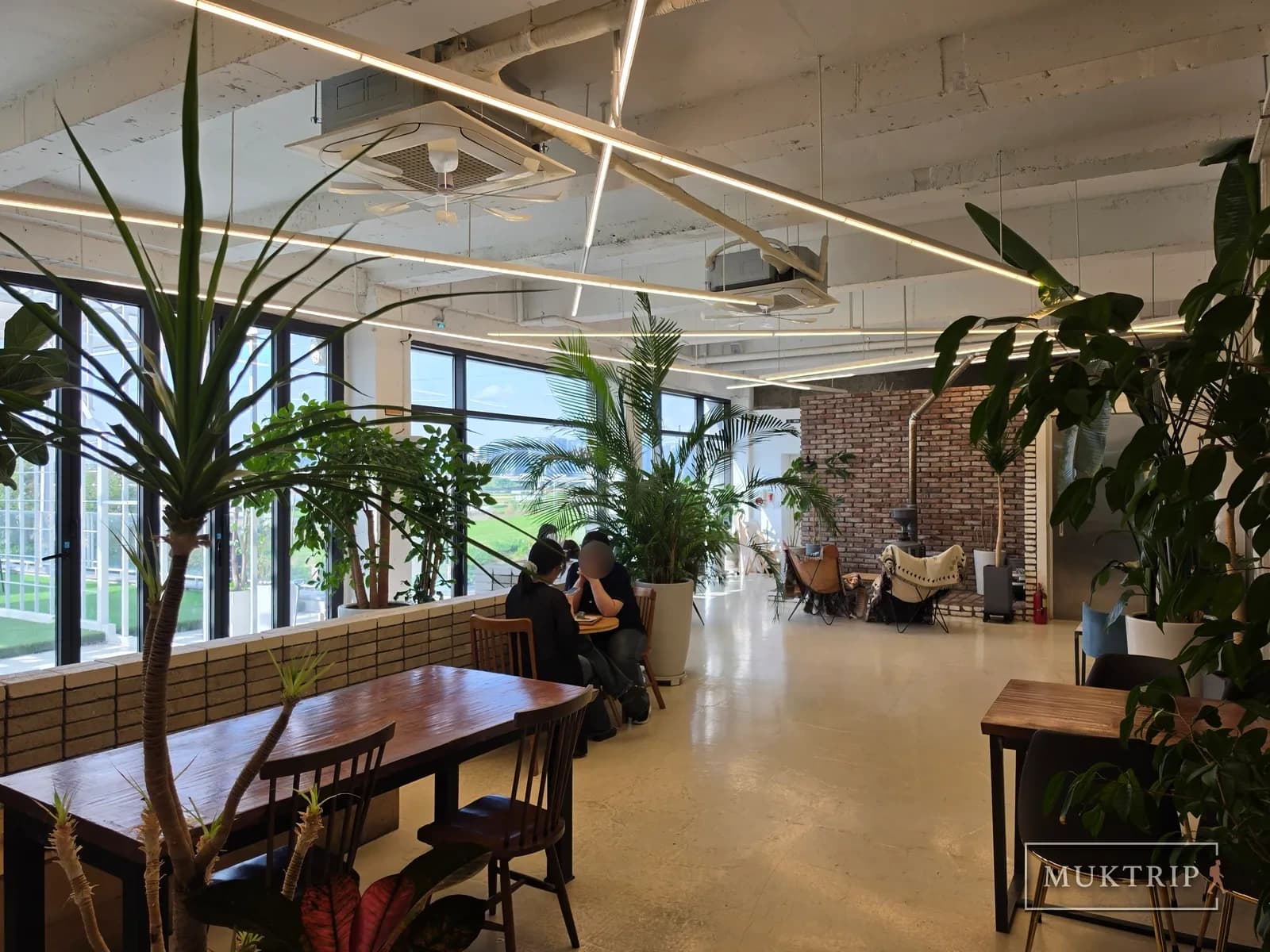 A spacious cafe hall combining floor-to-ceiling windows, green plants, and red brick walls