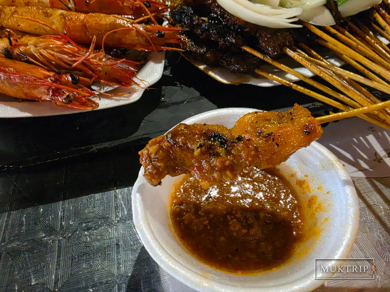 A grilled skewer being dipped into thick meat sauce, with prawns and satay skewers beside it