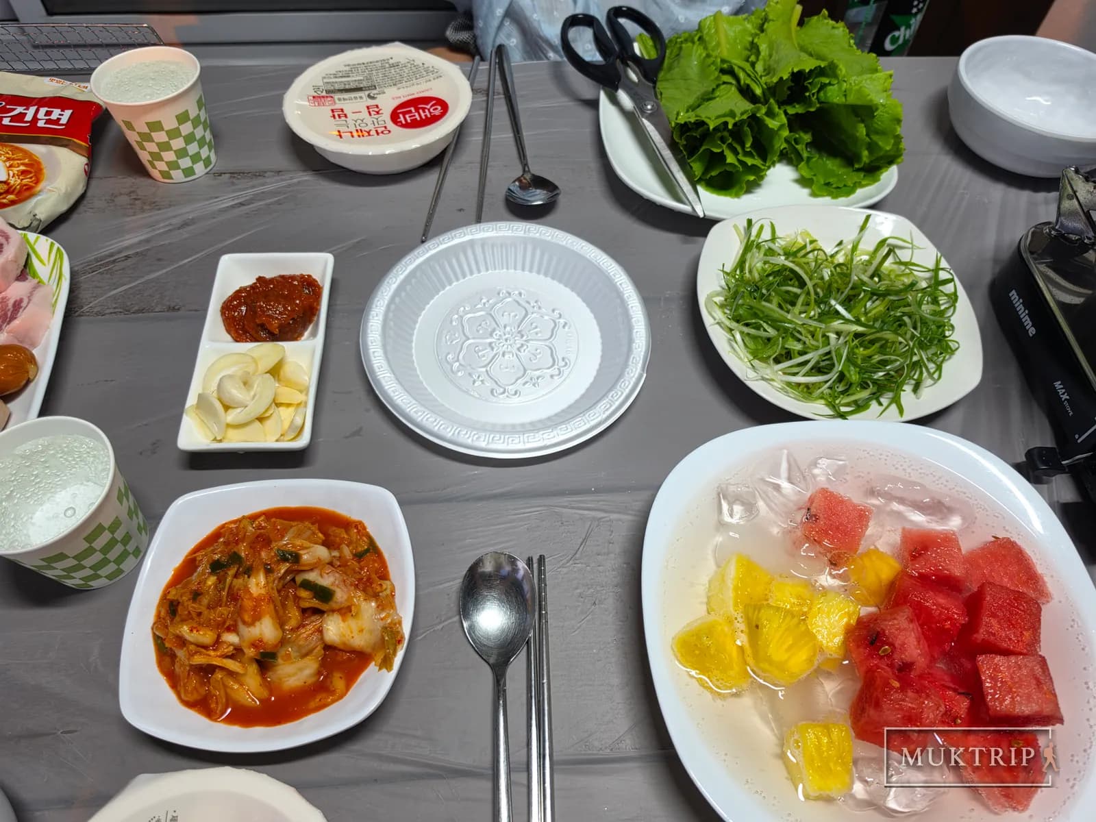 A barbecue side dish table set with lettuce, green onions, kimchi, and fruit punch