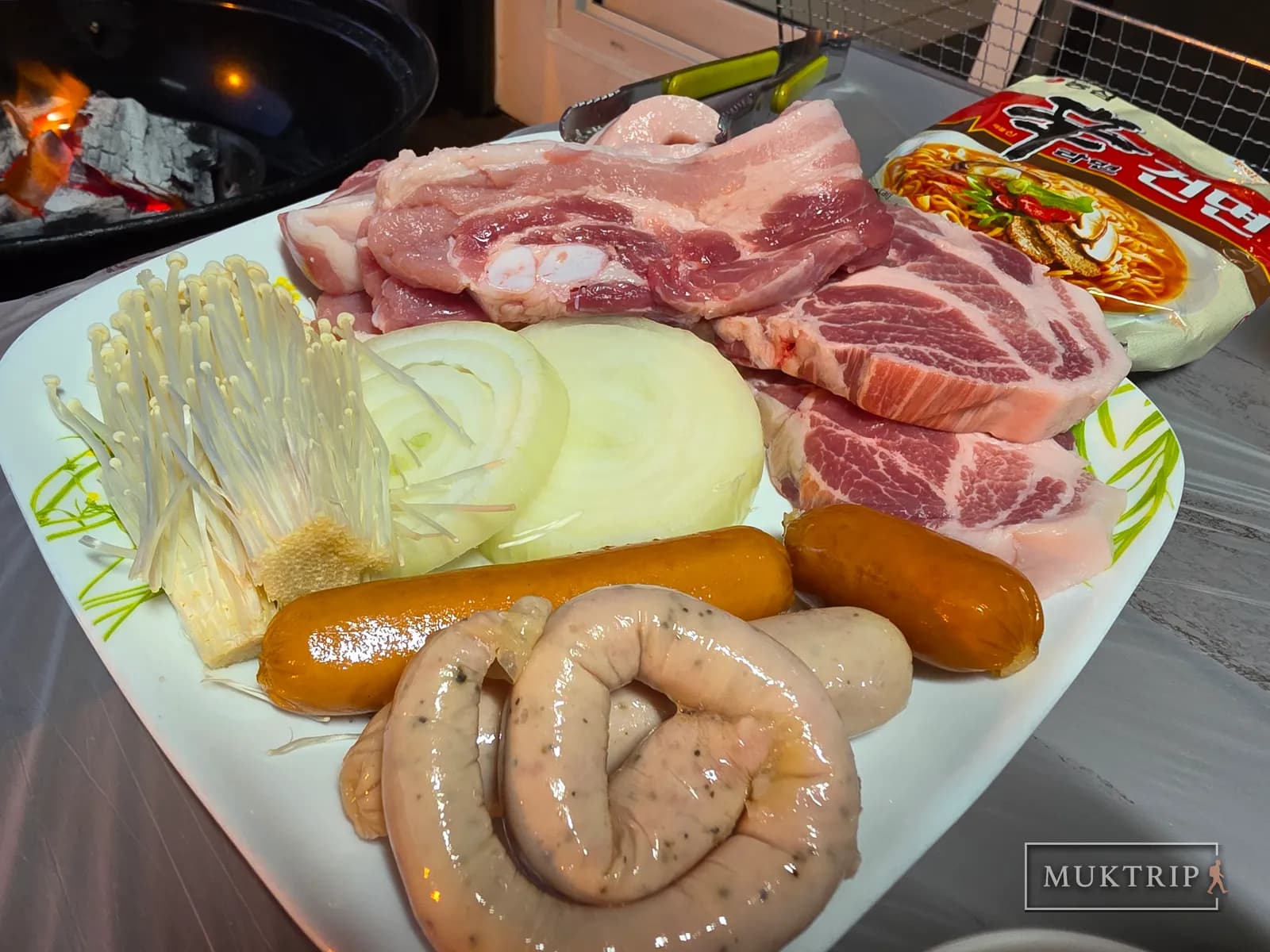 An assorted plate of pork belly, pork neck, sausage, and enoki mushrooms set in front of the charcoal fire