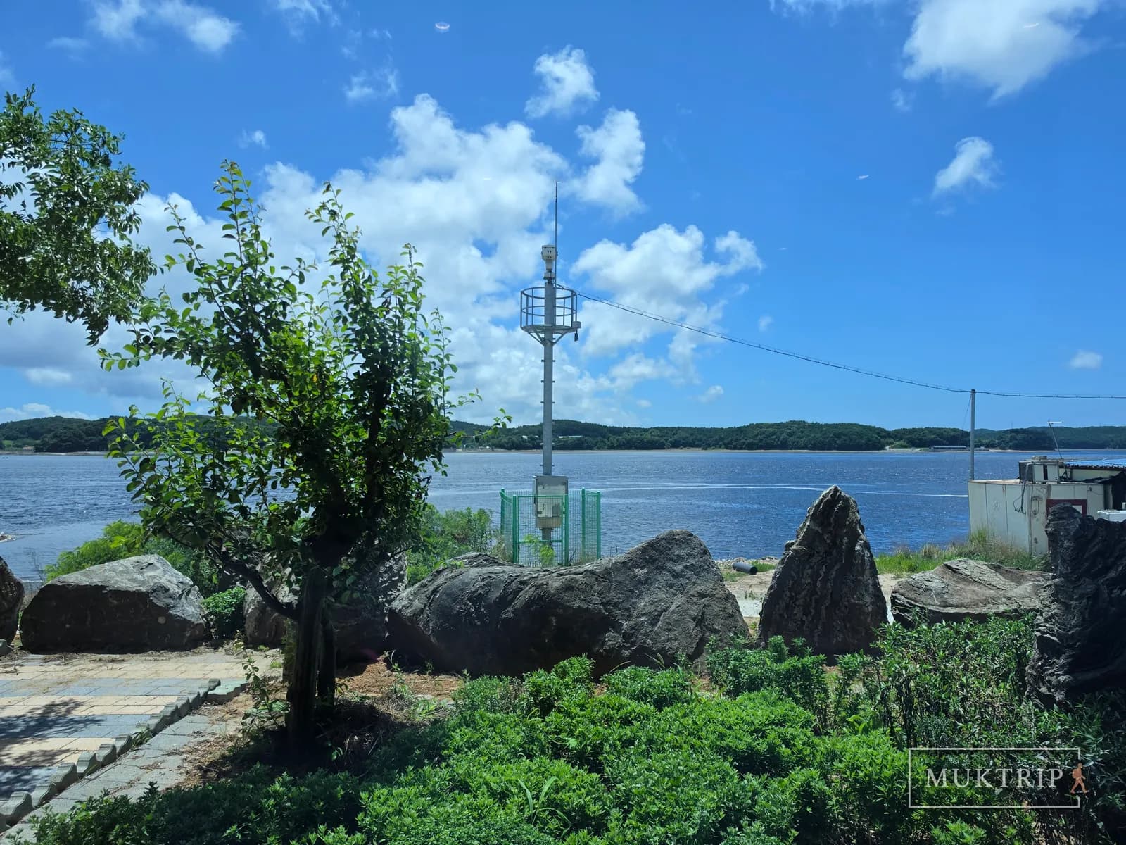 岩と木が調和した水辺の庭園の向こうに灯標が立っている